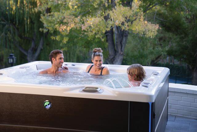  A couple and their young child enjoy a soak in a hot spring spa, with the hot tub's fountain feature turned on. Water cascades gracefully from the fountain, adding to the serene and playful atmosphere.