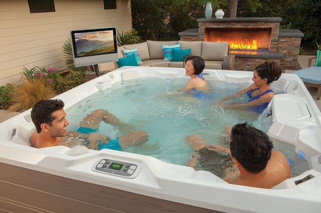 A group of friends relaxes in a luxurious Hot Spring Spa outdoor hot tub. The hot tub area is decked out with premium accessories: a large flat-screen TV mounted nearby and high-quality speakers strategically placed for optimal sound.