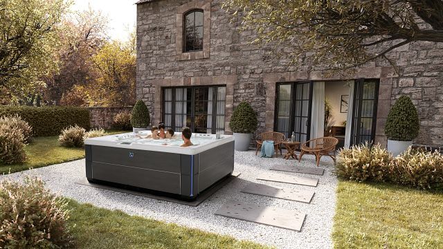 A family relaxes in their backyard hot spring spa, surrounded by the soothing, bubbling water. In the background, a beautiful stone house adds an elegant touch to the serene setting.