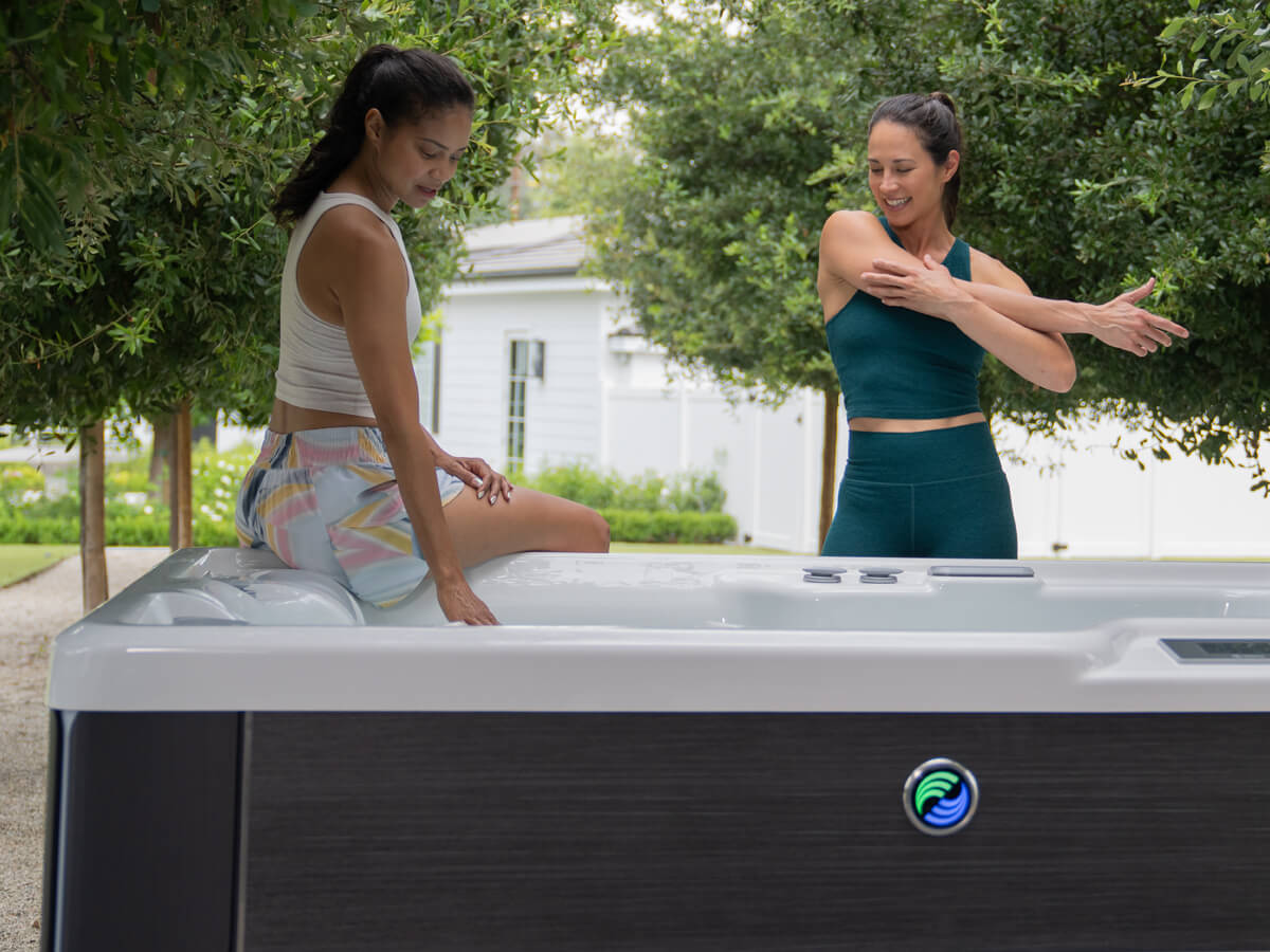 A woman sits at the edge of a Hot Spring Spa hot tub, touching the water, while another woman stands beside her, stretching her right arm after a workout.