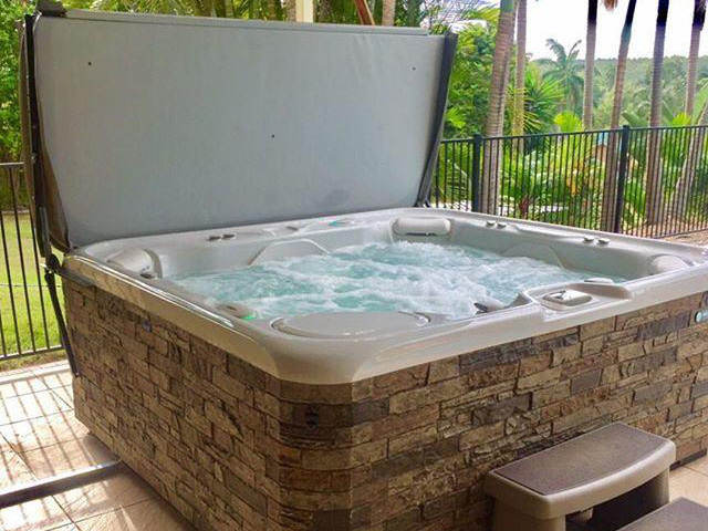 A luxurious outdoor hot tub with the cover open, surrounded by lush greenery. The hot tub is filled with bubbling water, ready for relaxation.