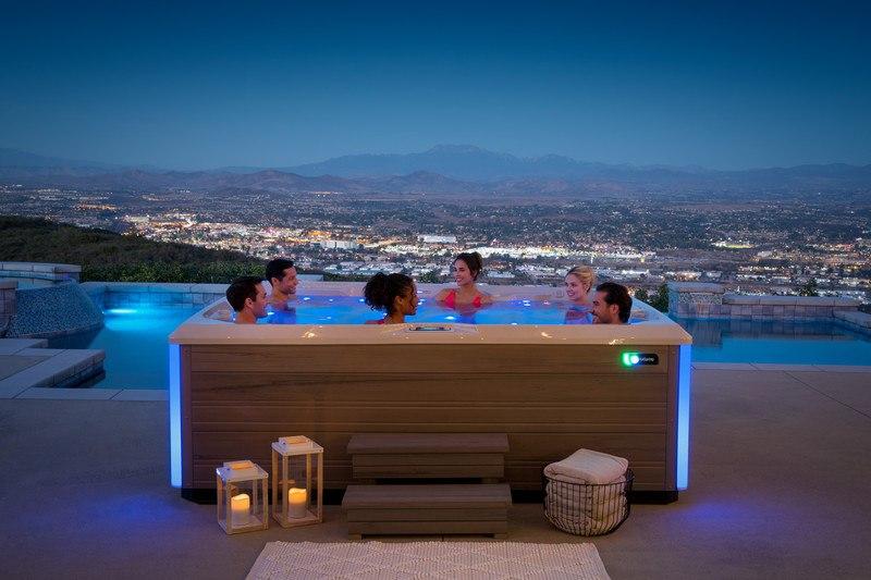 A group of six people relaxes in a Hot Spring Spas hot tub on a patio overlooking a city at dusk.