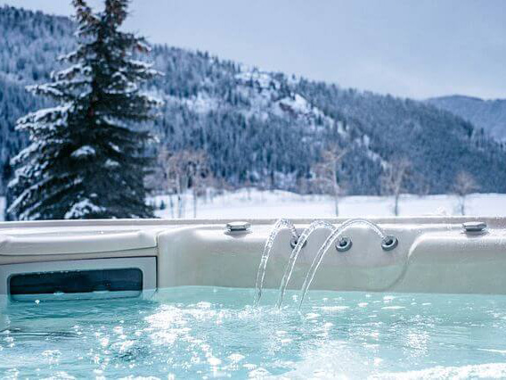 Close-up of a hot tub with flowing water jets, set against a snowy mountain landscape and pine trees in a serene winter setting.